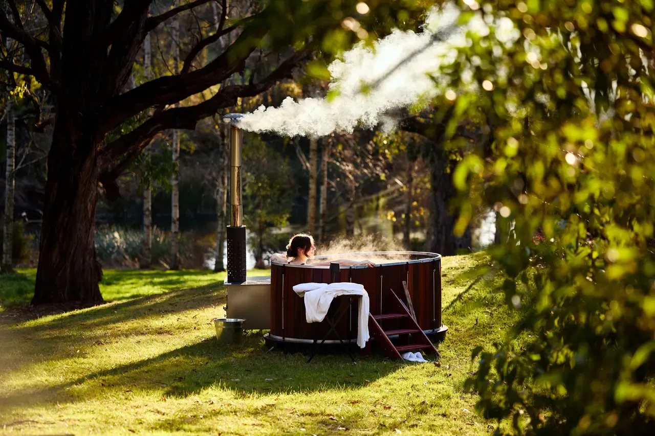 A guest soaks in a steaming wood-fired timber hot tub on a lawn beneath tall trees, a white towel draped over the steps