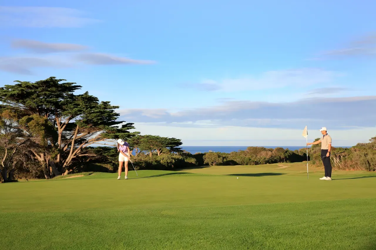 A golfer chips beside wind-shaped cypress trees at Flinders Golf Course while a partner holds the flag, ocean on the horizon