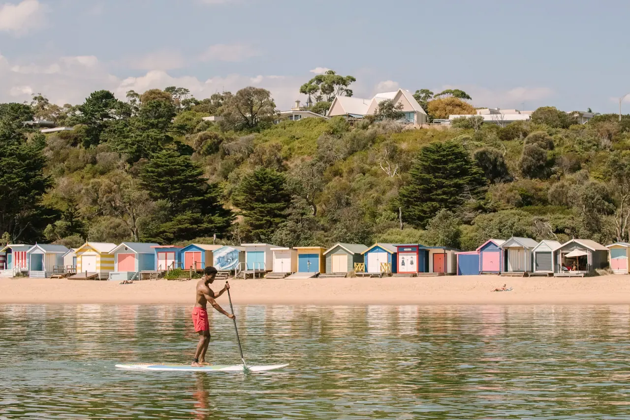 A stand-up paddleboarder glides across calm water past a row of colourful bathing boxes below a green hillside at Mornington