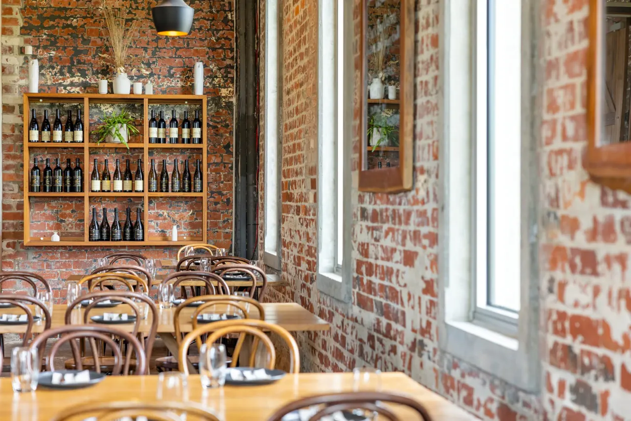Dining room at Trofeo Estate with exposed brick walls, bentwood chairs, set tables and a shelf of wine bottles