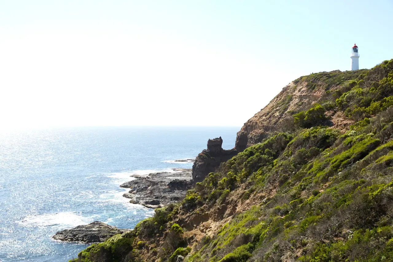 Scrubby cliffs at Cape Schanck falling to a rocky shore and sunlit sea, with the lighthouse on the headland above