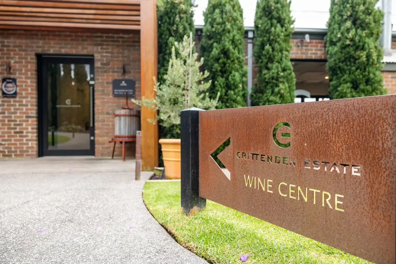 A rusted steel Crittenden Estate Wine Centre sign points along a path to the brick building's glass entrance