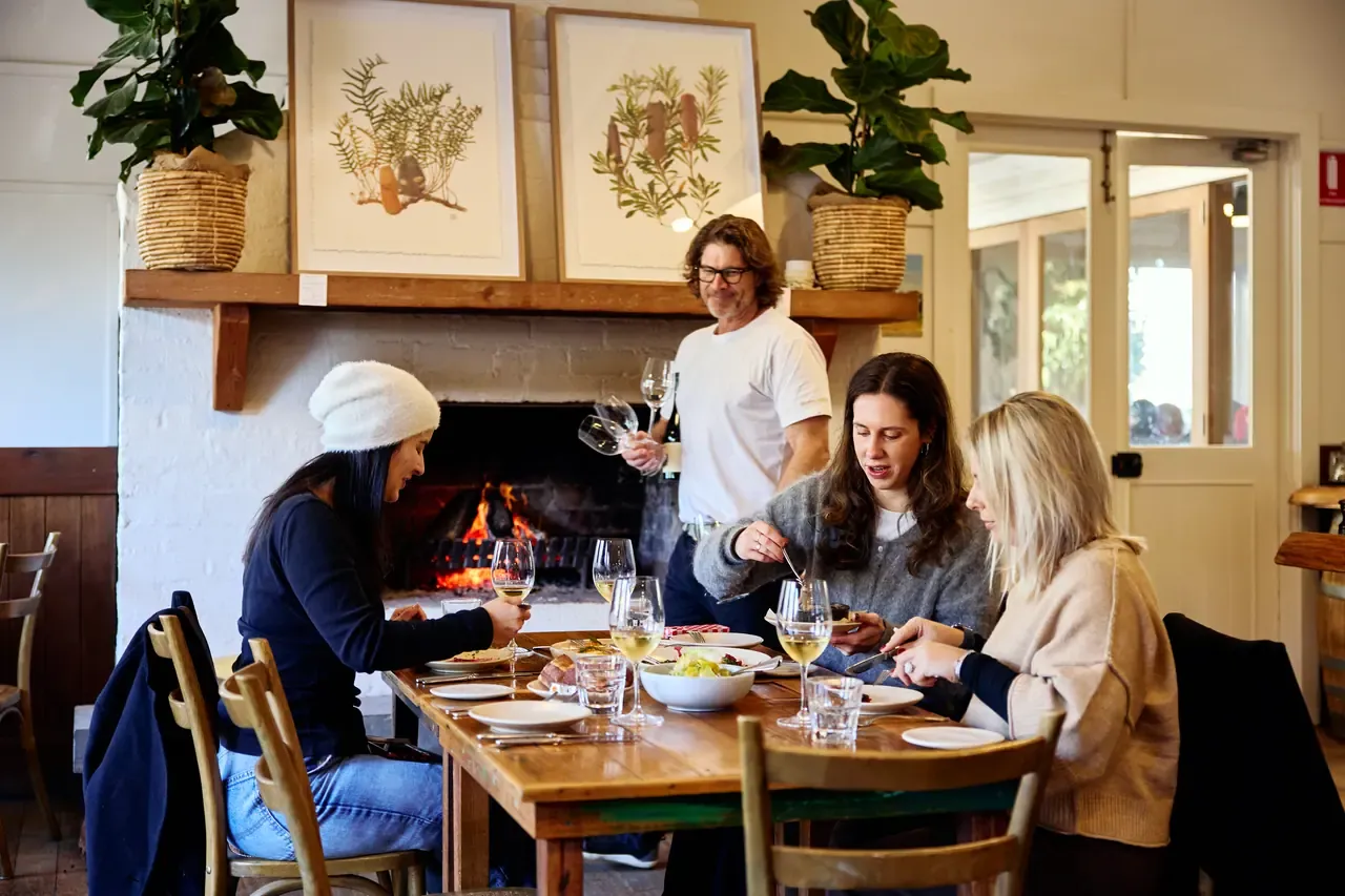 Three friends share plates and white wine at a table by the open fire while a waiter carries glasses at Merricks General Wine Store