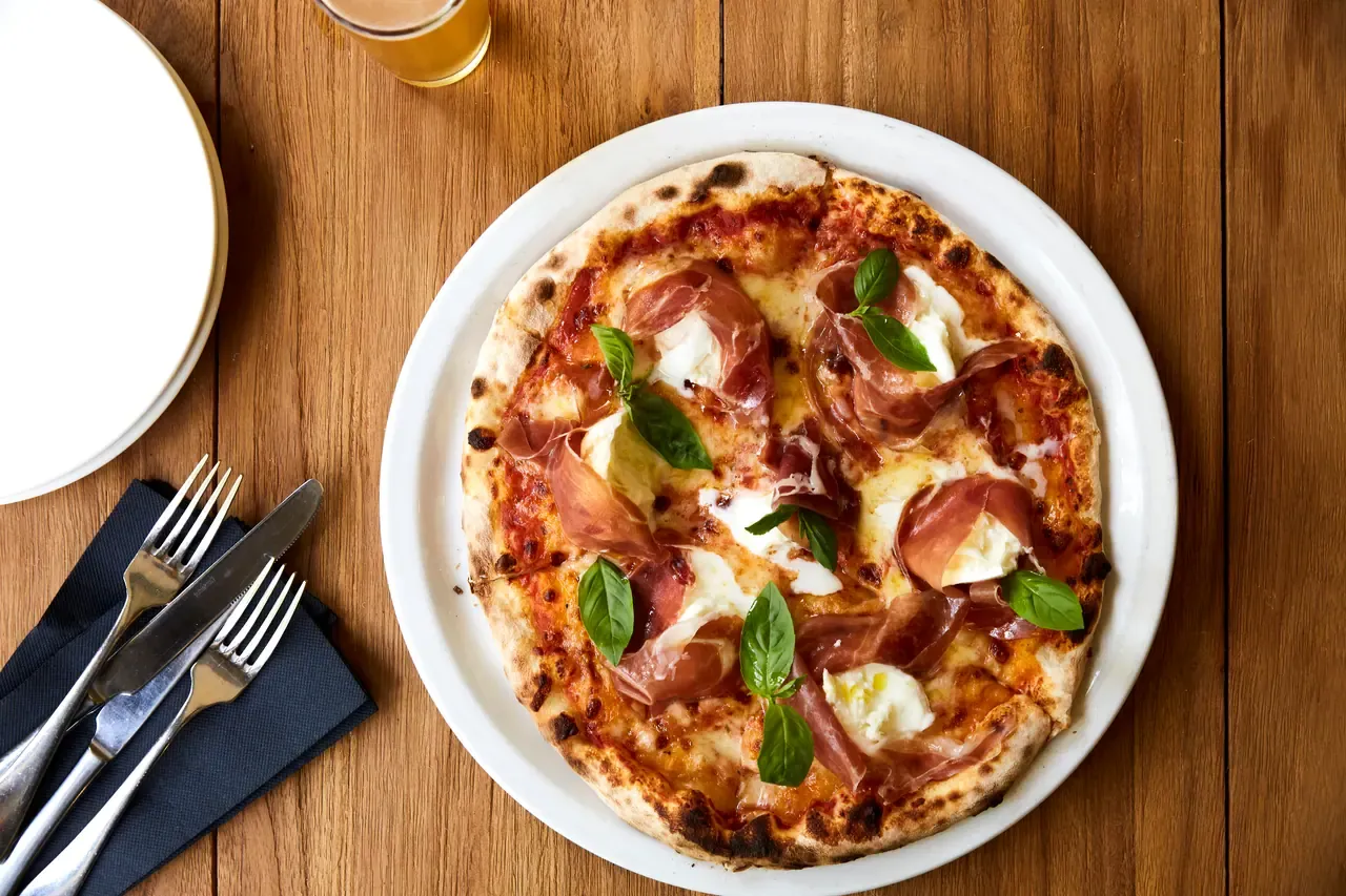 A wood-fired pizza topped with prosciutto, torn mozzarella and fresh basil on a white plate beside cutlery