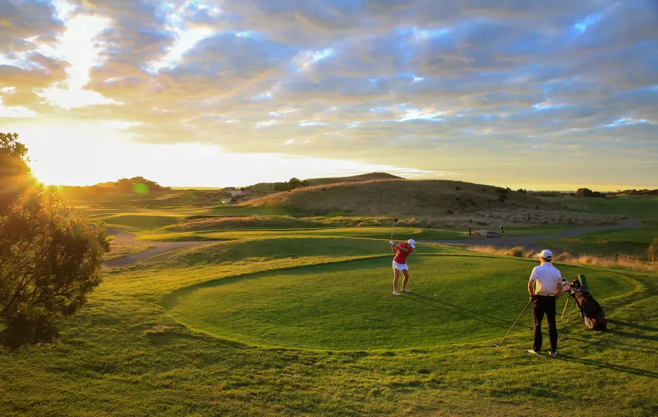 A golfer tees off at sunset at The Dunes Golf Links as a partner watches beside a golf bag, dunes glowing in the low sun