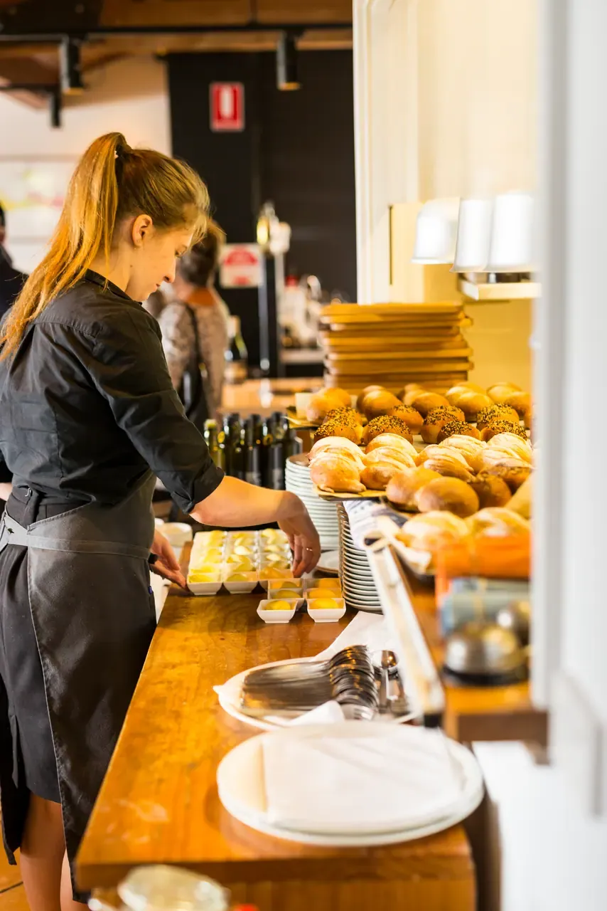 A staff member setting out small dishes of butter beside trays of bread rolls along a timber service counter