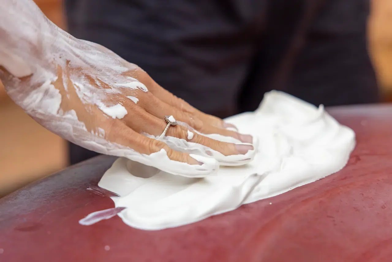 Close-up of a hand wearing a ring spreading thick white cream across a smooth dark red surface, likely part of a spa treatment