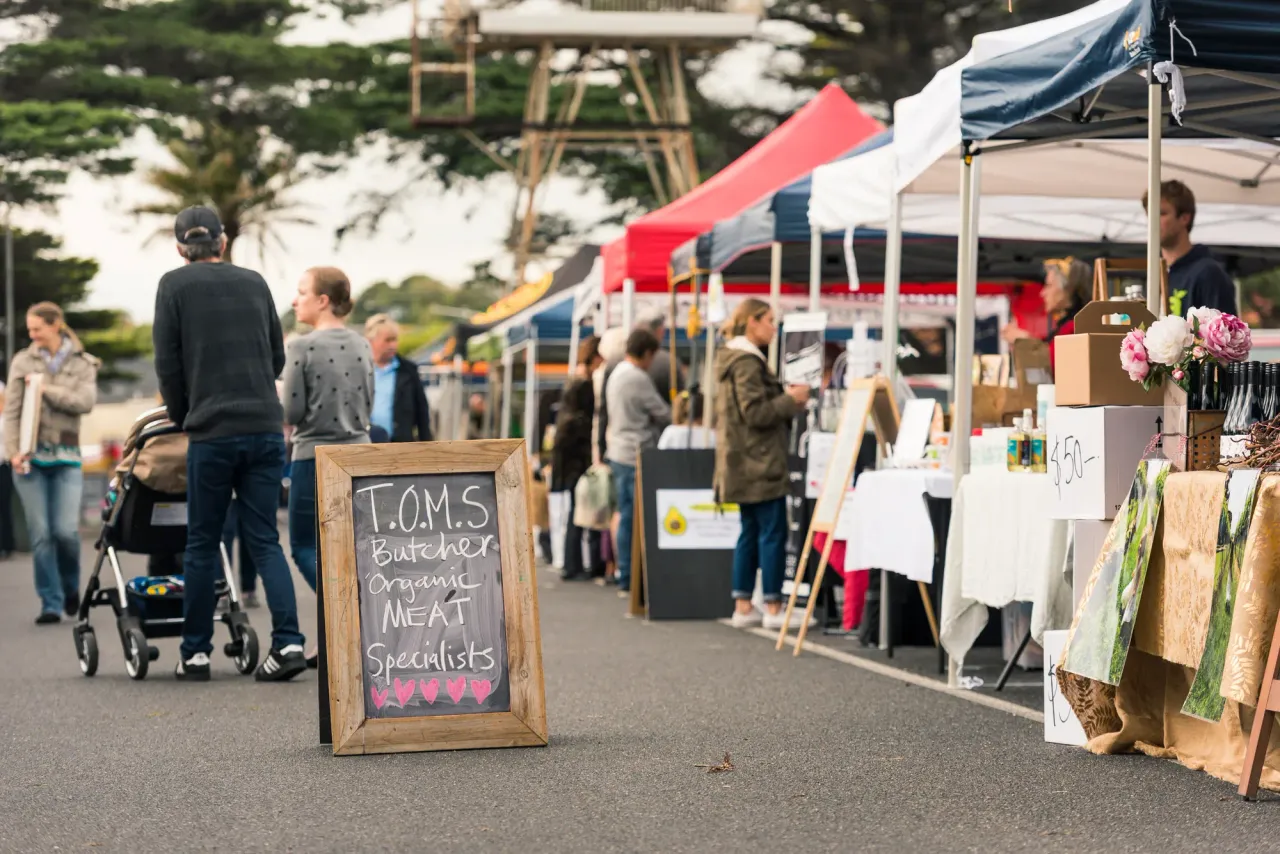 Shoppers stroll past white and red marquees and a butcher's chalkboard sign at Mornington Farmers Market