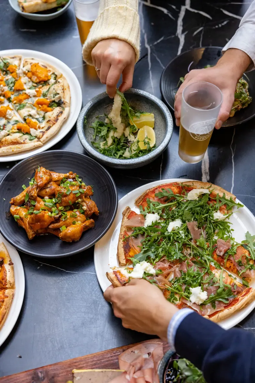Overhead view of hands reaching for pizzas, chicken wings and a salad on a dark marble table, with glasses of beer