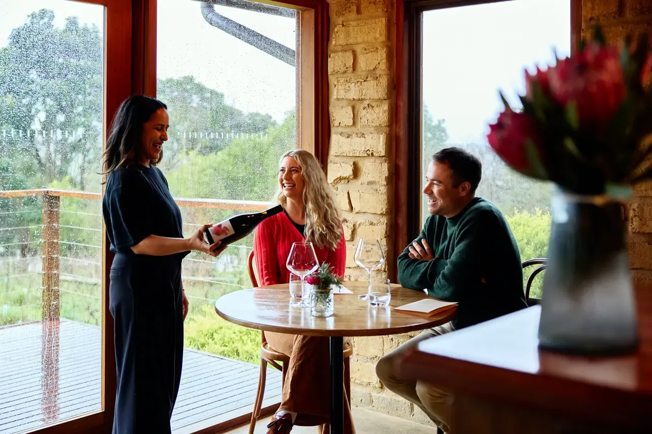 A host presenting a bottle of wine to two smiling guests at a window table in the Avani Winery cellar door