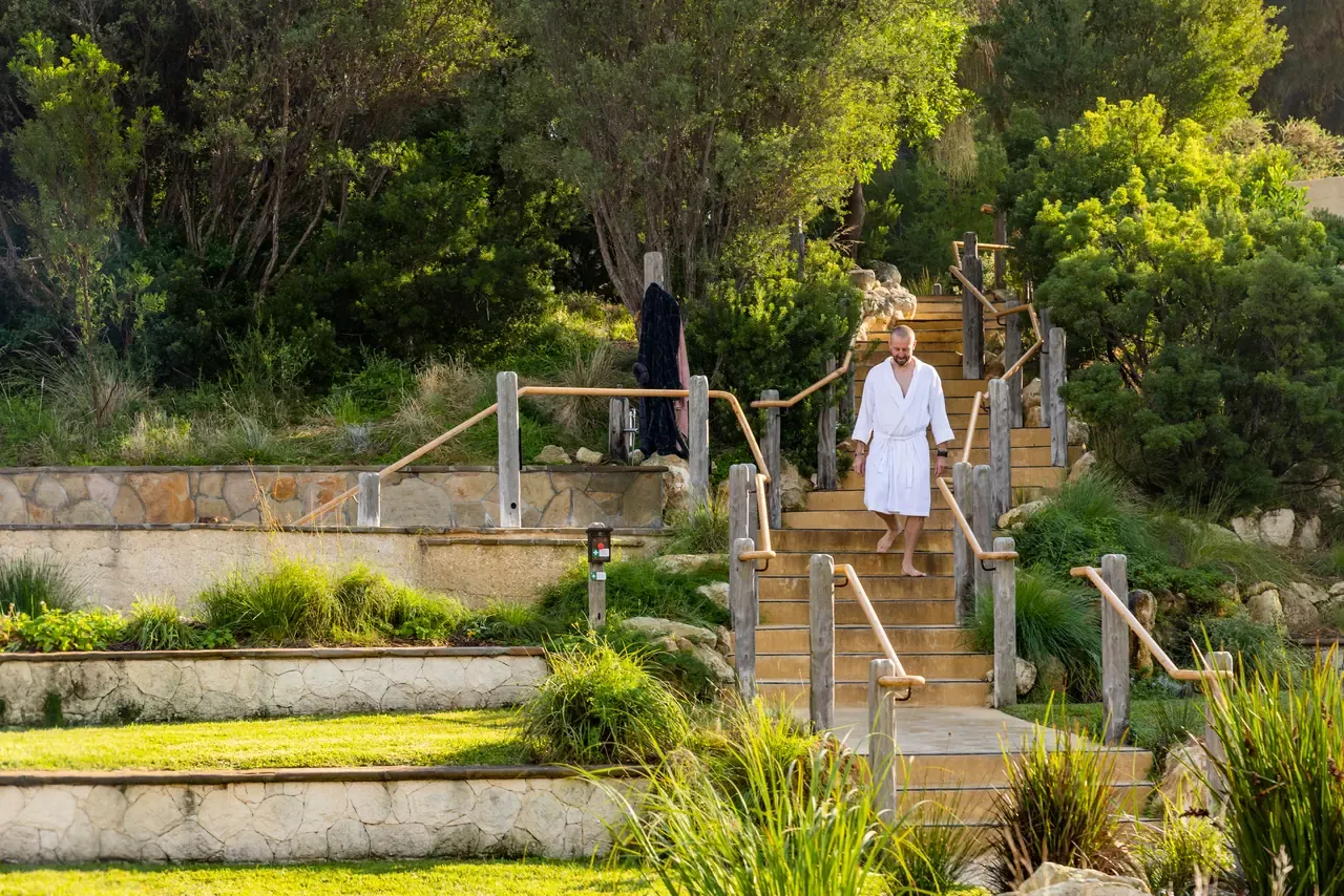 Guest in a white robe walks down timber stairs with rope handrails, past stone-walled terraces and native garden at Peninsula Hot Springs