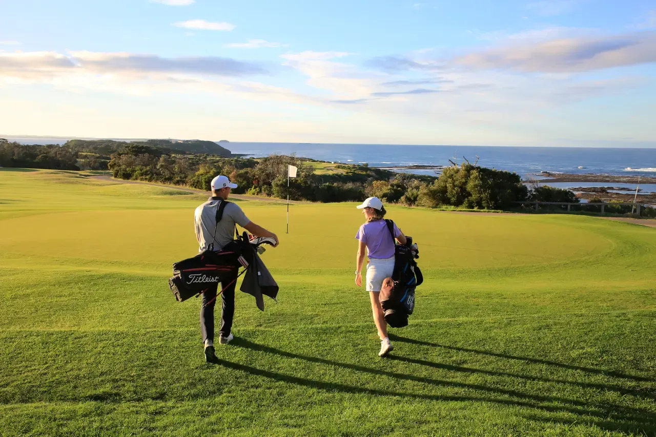 Two golfers with bags walk across a sunlit green towards a flag, with the ocean and coastal scrub beyond at Flinders Golf Course.