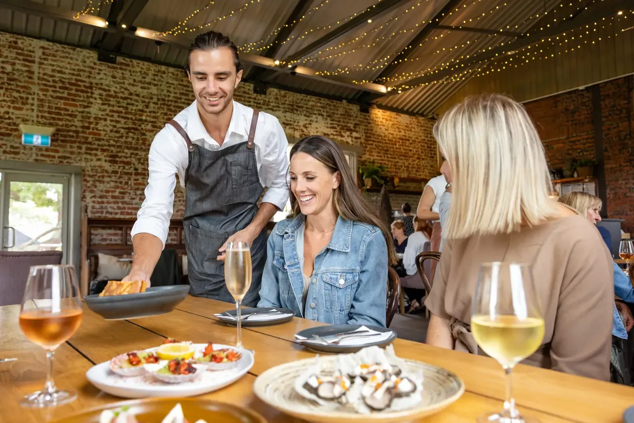 A smiling waiter serves shared plates and wine to guests at a long table under festoon lights at Trofeo Estate