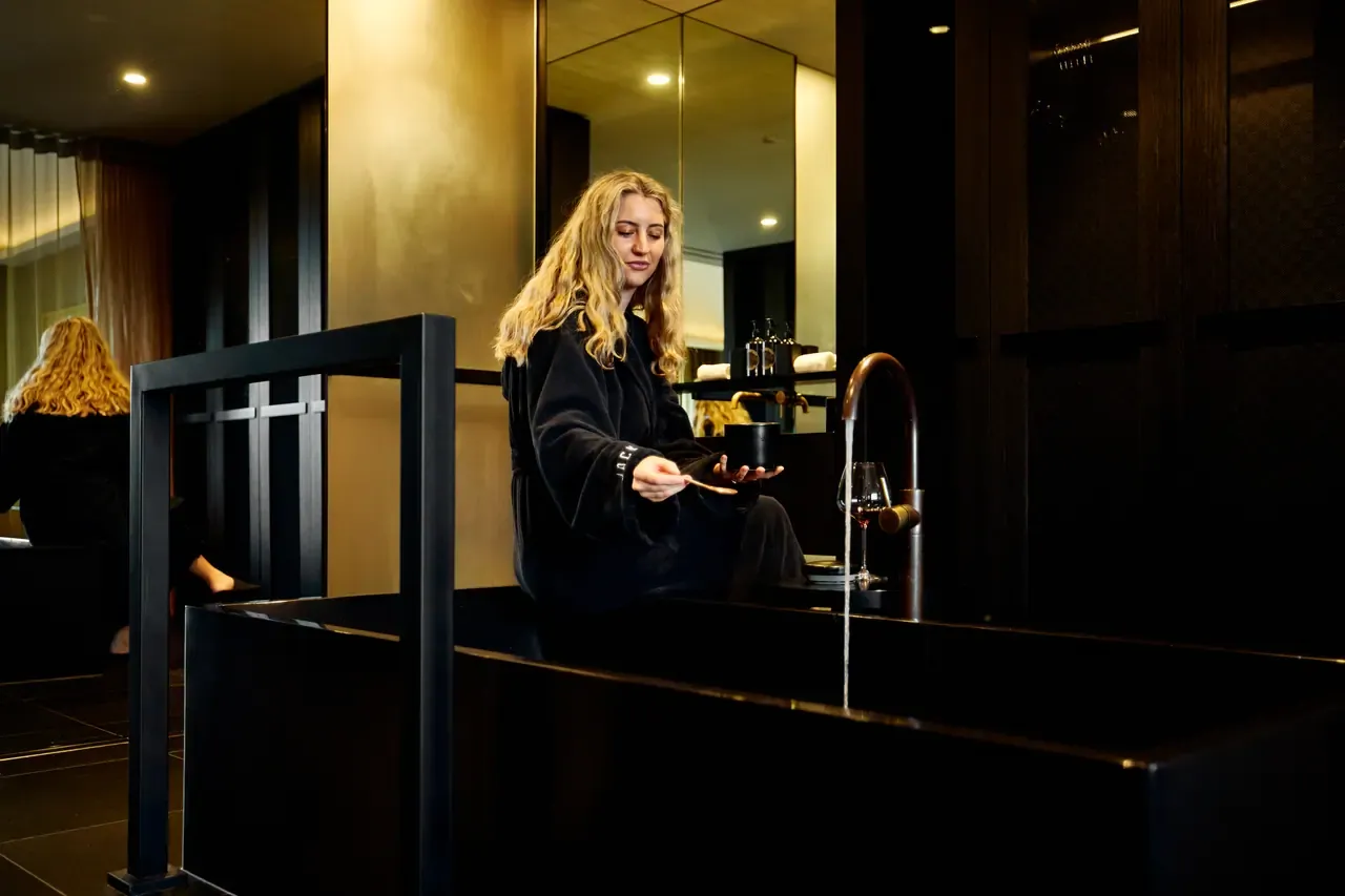 A guest in a dark robe sits beside a deep black bath as water runs from a curved brass tap in a Jackalope Hotel suite