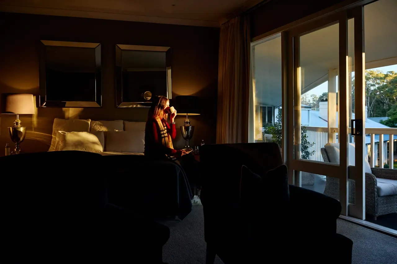 A guest sips from a mug on the edge of the bed as low sun lights a darkened room with a balcony at Lindenderry
