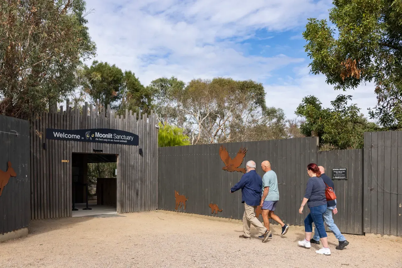 Visitors walk across a gravel forecourt towards the timber-fenced entrance and welcome sign of Moonlit Sanctuary