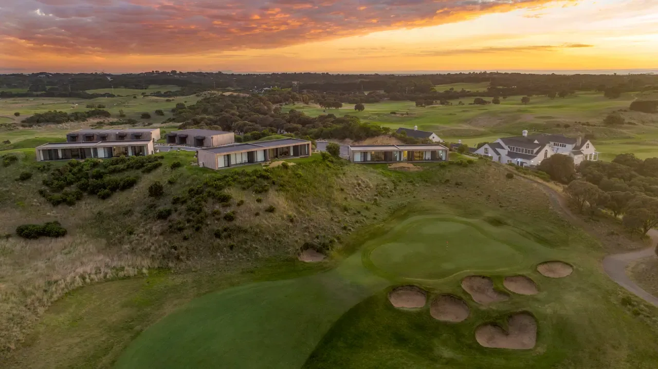 Aerial view of a links golf green ringed by bunkers, with low modern accommodation and a homestead beneath a fiery sunset sky.