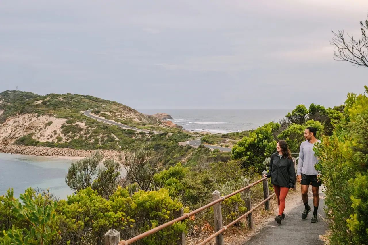 Two walkers on a railed path through coastal scrub at Point Nepean, the winding headland road and sea stretching beyond