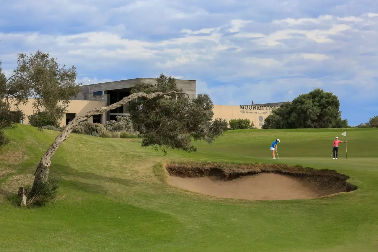 Golfers on a green beside a bunker and a leaning tree, with the Moonah Links clubhouse and signage behind.