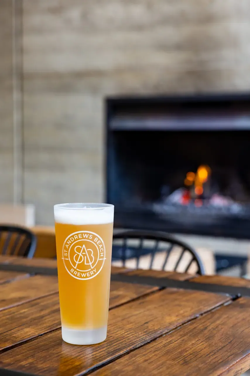 A glass of pale beer on a timber table at St Andrews Beach Brewery, with a fireplace glowing in the background