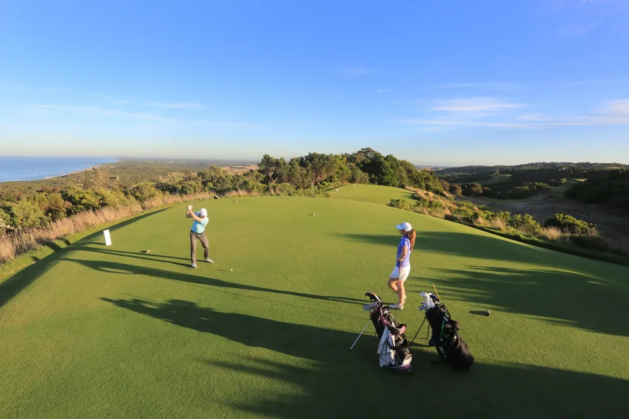 A golfer swings on a raised tee at The National Golf Club as a partner waits by two bags, with coastal bush and the sea beyond