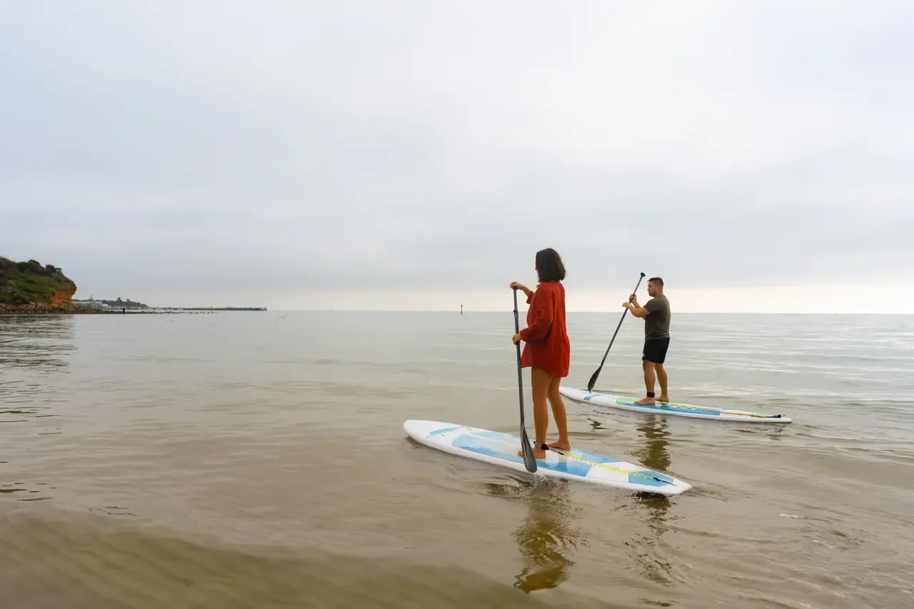 Two stand-up paddleboarders glide across calm grey water off Mills Beach under an overcast sky