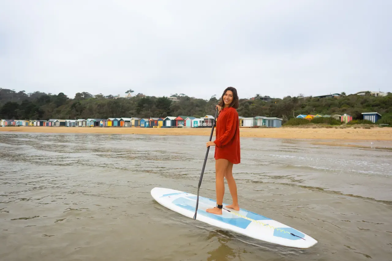 A paddleboarder in a red shirt stands on her board in the shallows at Mills Beach, with bathing boxes along the shore