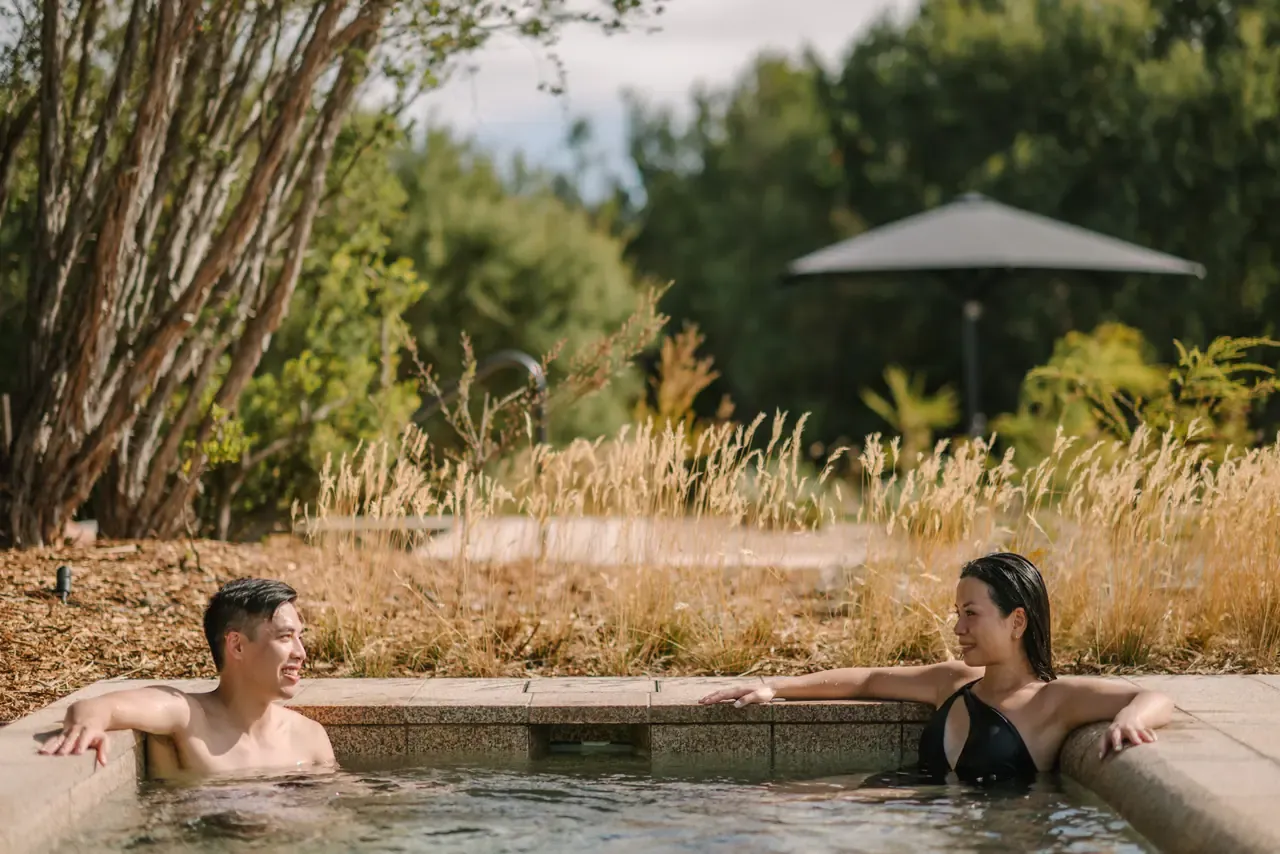 Two bathers lean on the edge of an outdoor pool with golden grasses, native trees and a shade umbrella behind them