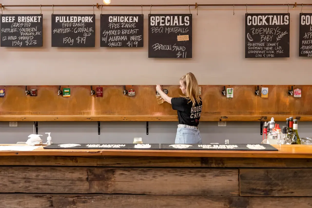 A staff member works at the long timber bar at Red Gum BBQ under chalkboard menus for brisket and pulled pork