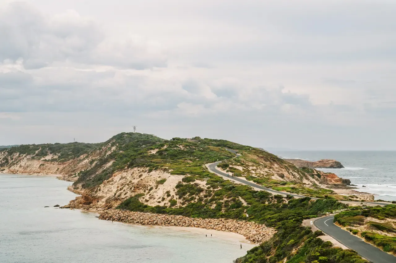A winding road runs along a scrub-covered headland at Point Nepean, with a rock seawall and calm bay water under grey cloud