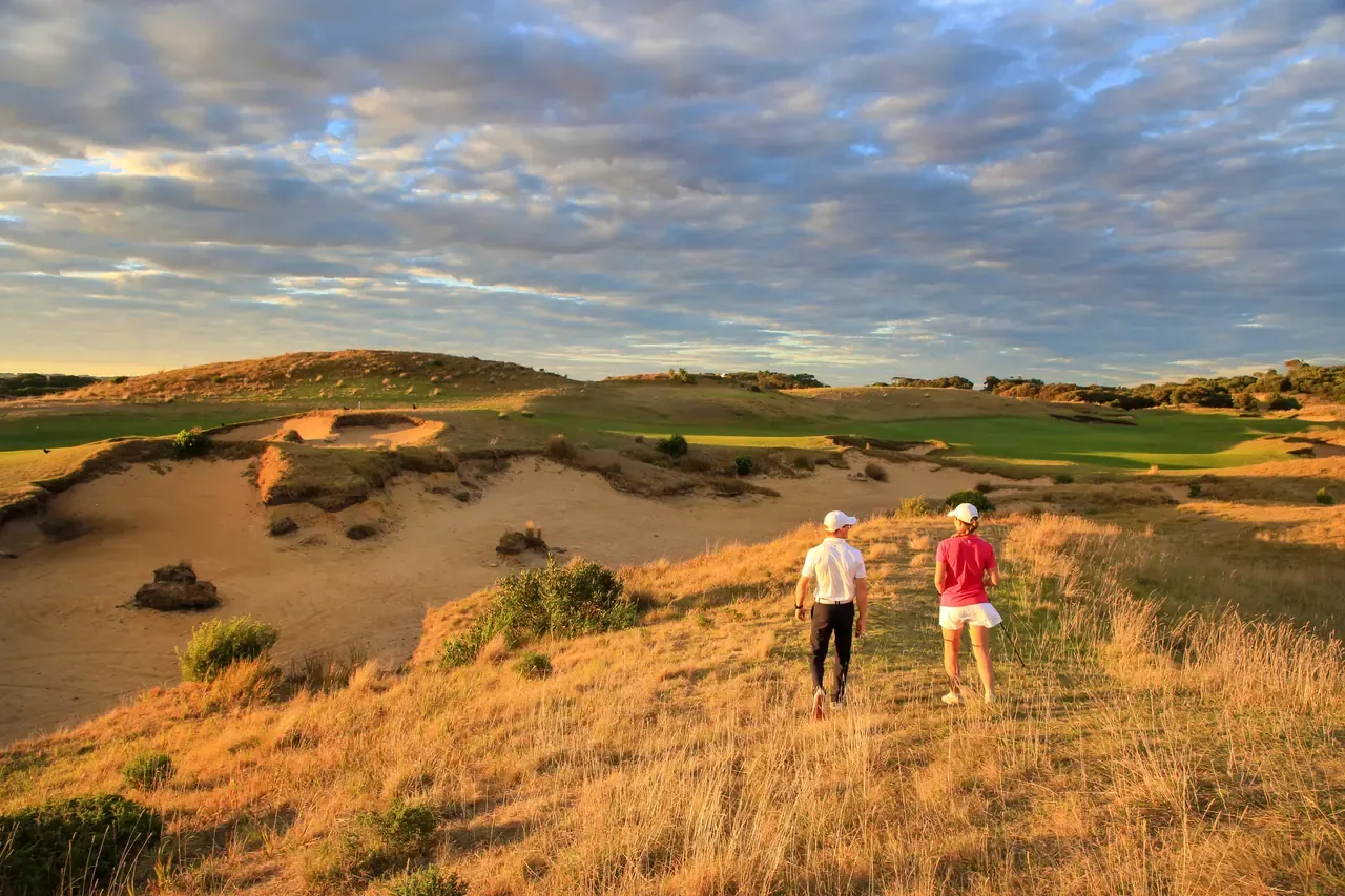 Two golfers walk through golden dune grass beside a large sandy waste area at The Dunes Golf Links under a streaked evening sky