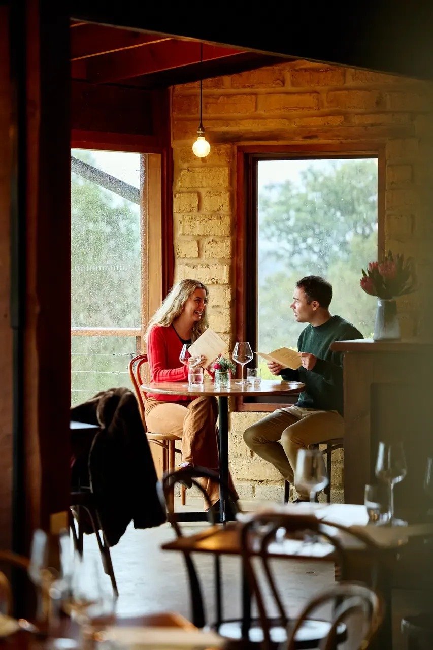 Two guests reading menus at a table by rain-streaked windows in the mudbrick dining room at Avani Winery