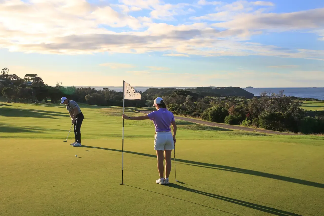 One golfer putts while another holds the flag on a green at Flinders Golf Course, with headland and sea behind in soft evening light.