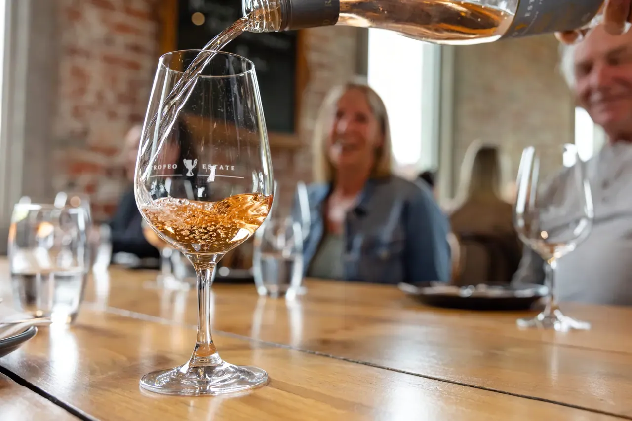 Rosé being poured into a Trofeo Estate branded glass on a timber table, with smiling guests blurred in the background