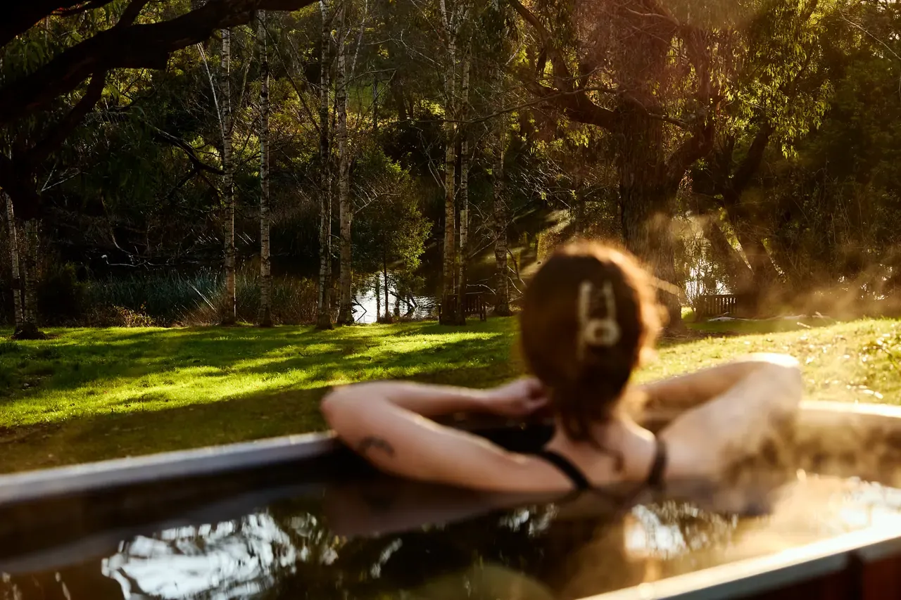 A bather relaxes in a steaming outdoor tub facing a lawn, lake and trees in late light at Lindenderry at Red Hill