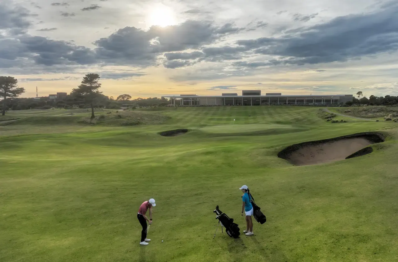 Two golfers play a chip shot beside a bunker on a rolling fairway at Moonah Links, with the clubhouse under a moody evening sky.
