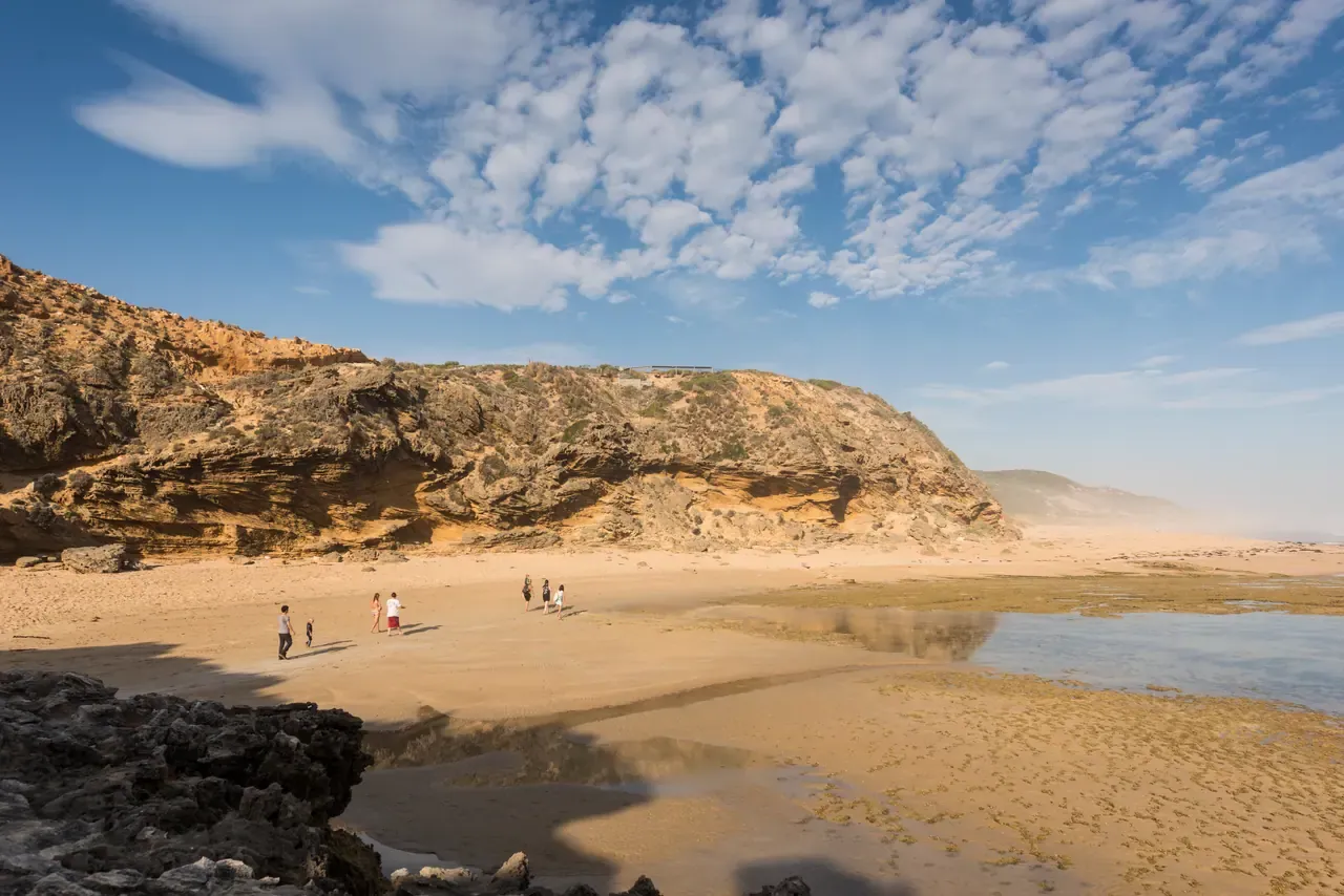A few people walk across a wide sandy beach at low tide beneath golden limestone cliffs on the Portsea ocean coast