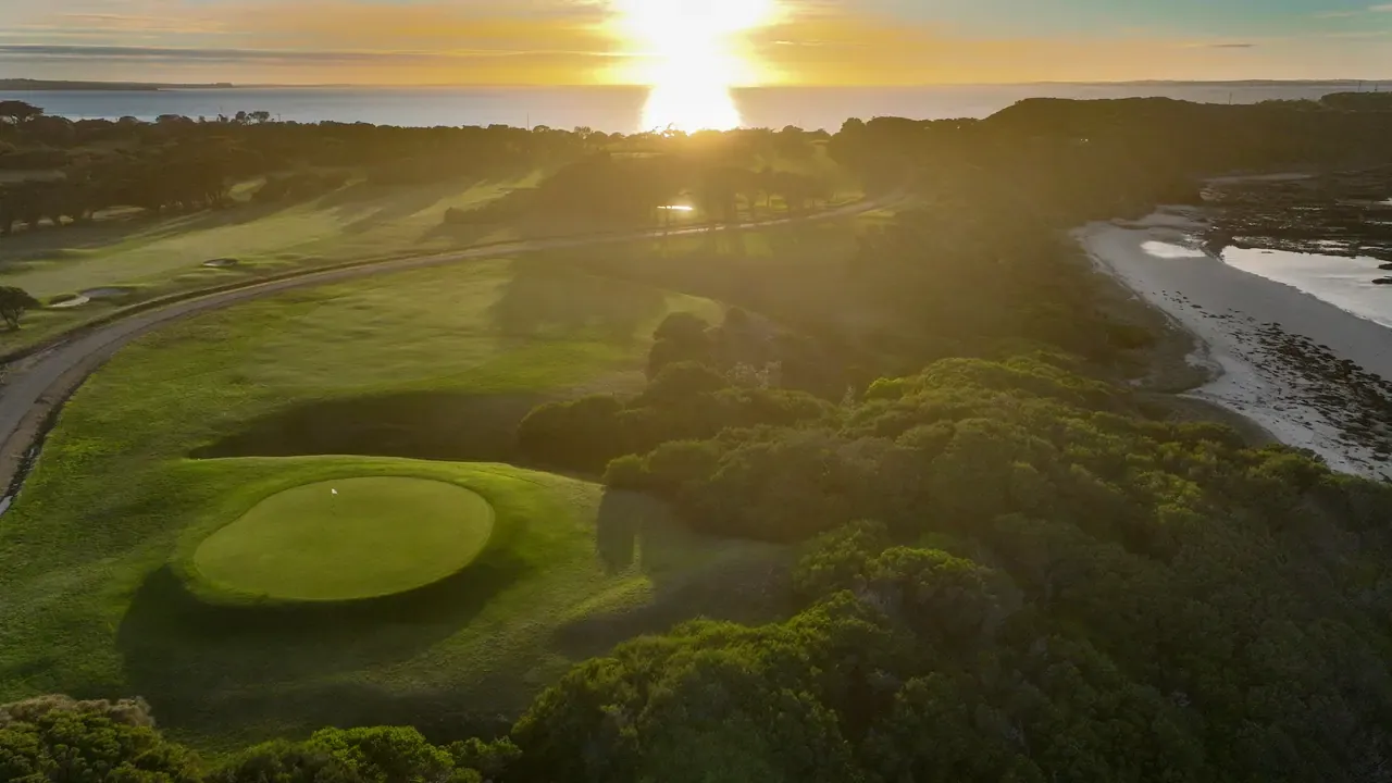 Aerial view of a Flinders Golf Course green ringed by coastal scrub, with low sun glowing over the water and a beach at right.
