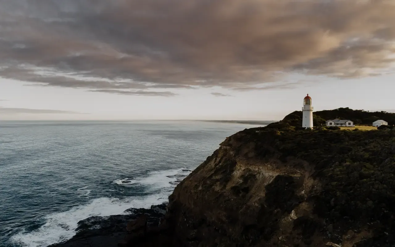 Aerial view of Cape Schanck Lighthouse and its cottages on a scrub-covered clifftop above surf, under heavy cloud at low sun