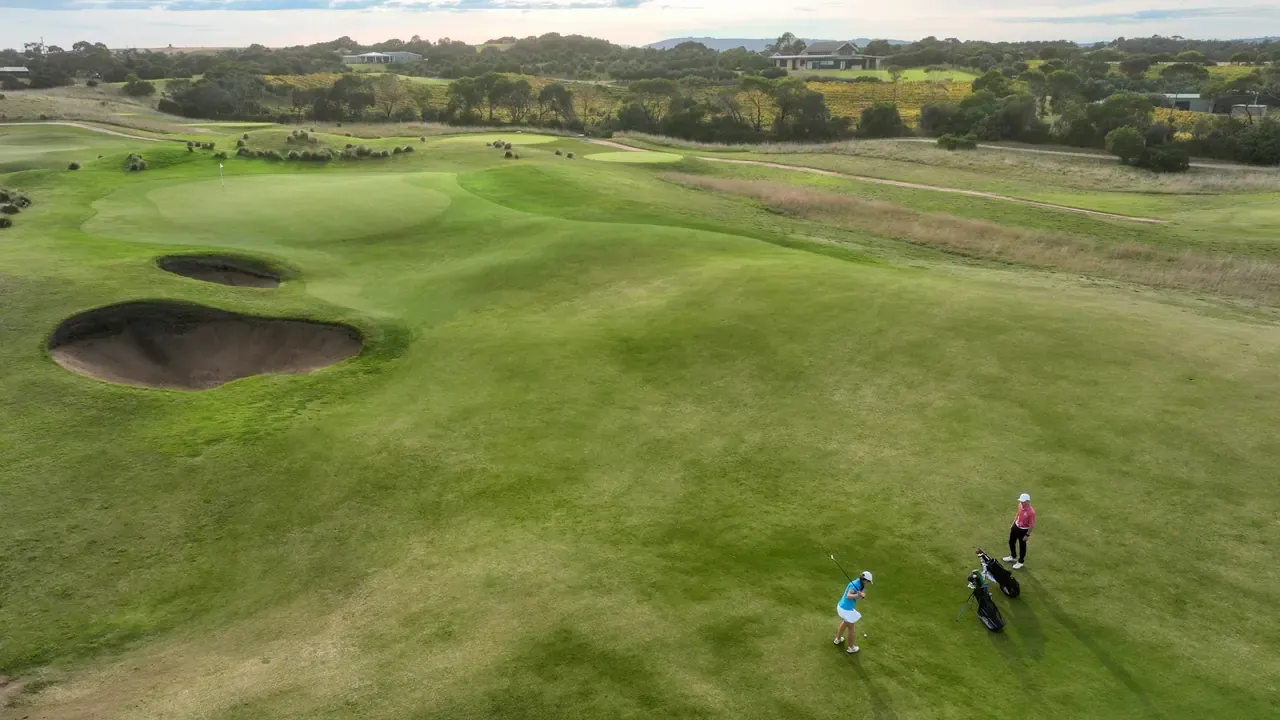 Aerial view of two golfers playing towards greens and deep bunkers at Moonah Links Golf Course, Fingal, with rural land beyond.