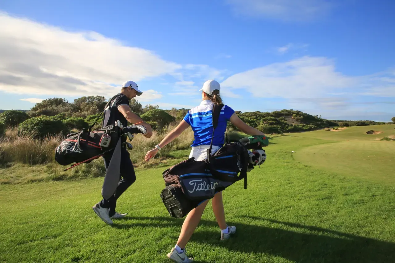 Two golfers carry their bags across the fairway at St Andrews Beach Golf Course under a blue sky with scrubby dunes behind