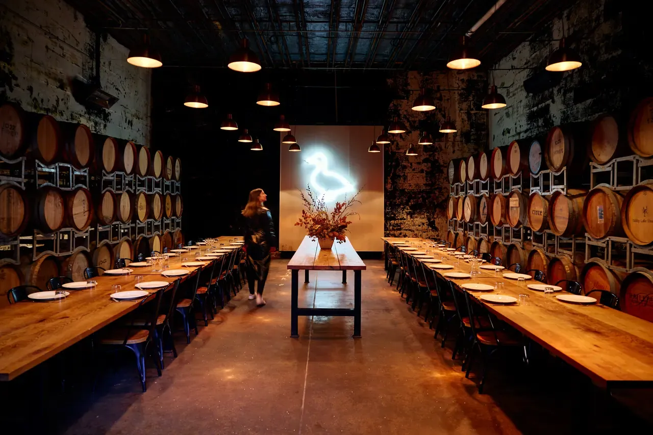 Long dining tables set between walls of stacked wine barrels under a neon swan sign at The Epicurean, Red Hill