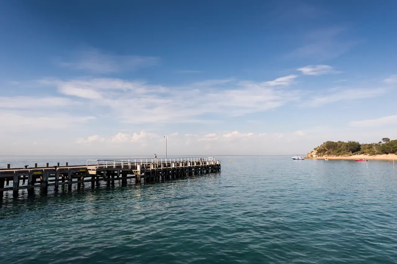 The timber Portsea Pier reaching into calm blue water, with moored boats and a bush-covered point beyond
