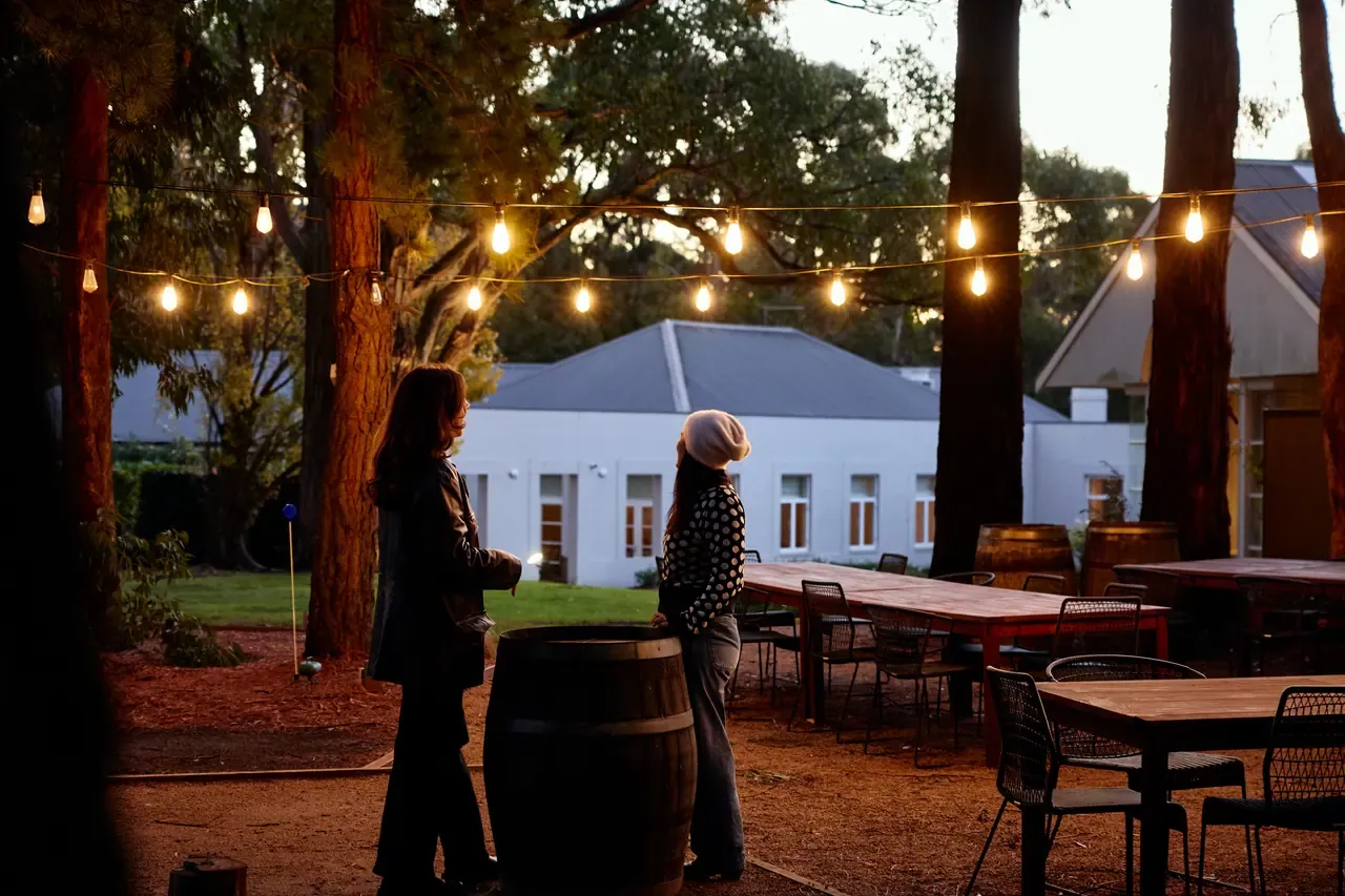 Two guests stand beside a wine barrel under festoon lights near outdoor tables and the white homestead at Lindenderry at dusk