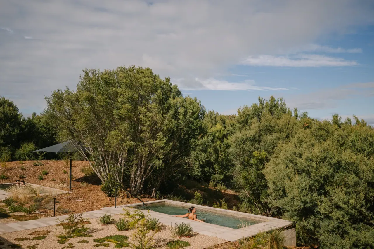 Bather in a rectangular outdoor pool edged with pale paving, set among dense coastal tea trees under a partly cloudy sky