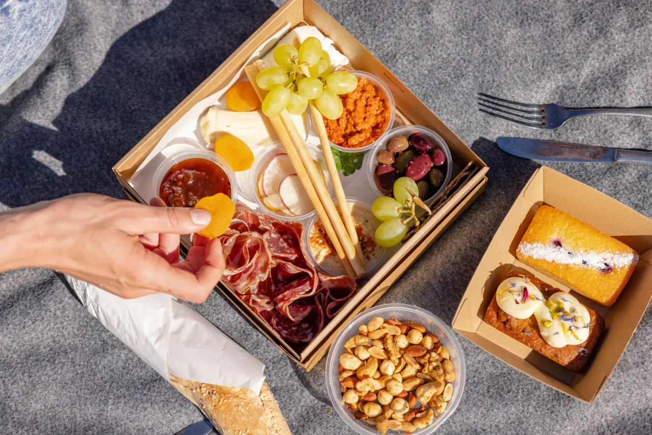 A picnic box of cured meats, cheese, grapes, dips and olives on a rug, with nuts and a slice of cake alongside