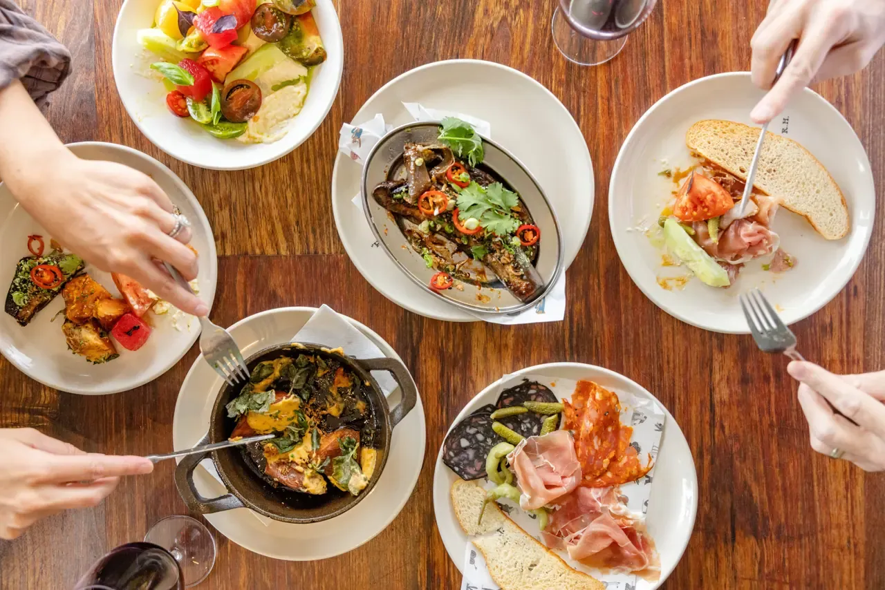 Overhead view of shared plates on a timber table: cured meats, tomato salad, a mushroom dish and a cast-iron pot, with diners' hands