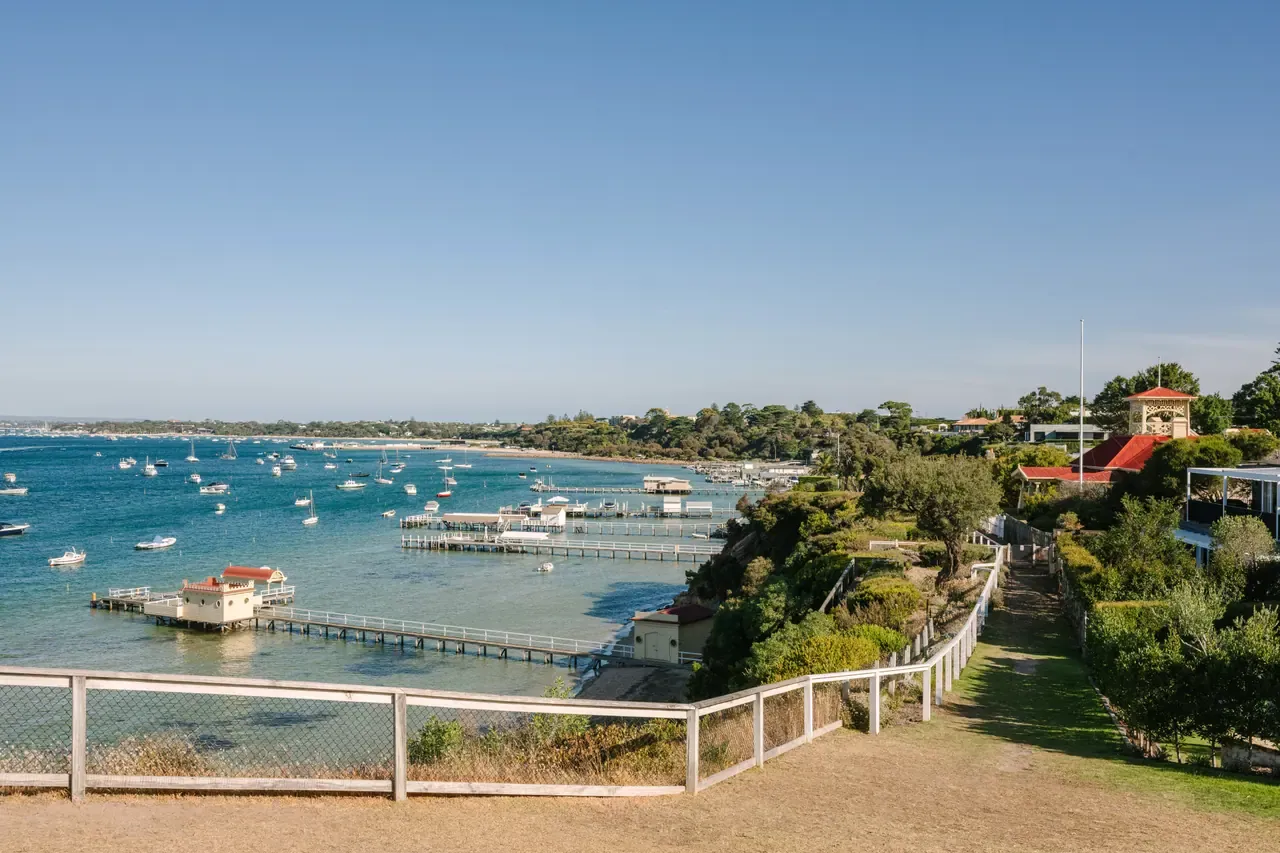 A fenced clifftop path on Millionaire's Walk looks over boat sheds, jetties and moored boats in a calm blue bay