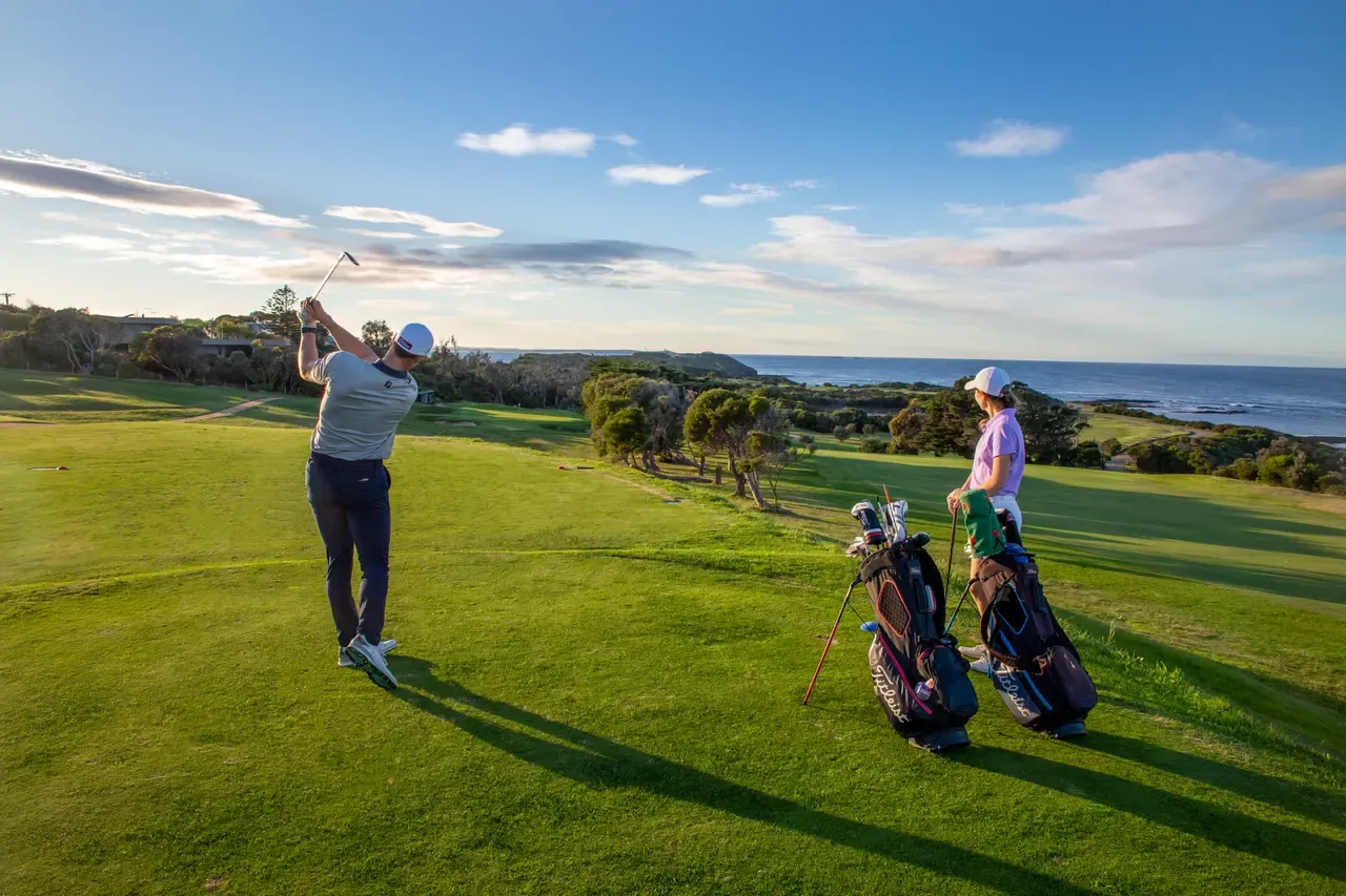 A golfer tees off on a coastal hole at Flinders Golf Course while a partner waits by the bags, sea and headland beyond