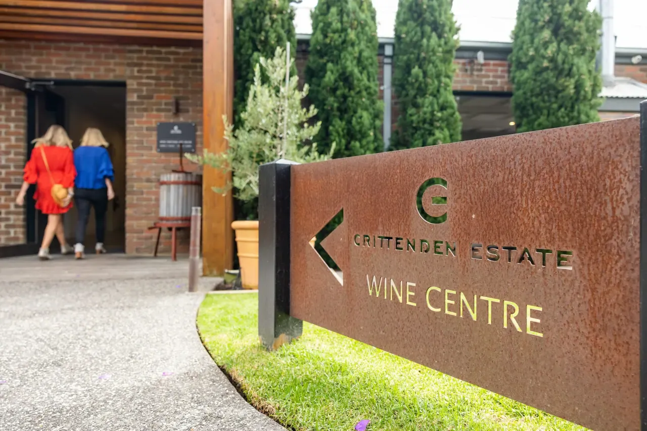 Two visitors walk into the Crittenden Estate wine centre past a rusted steel sign and tall cypress trees